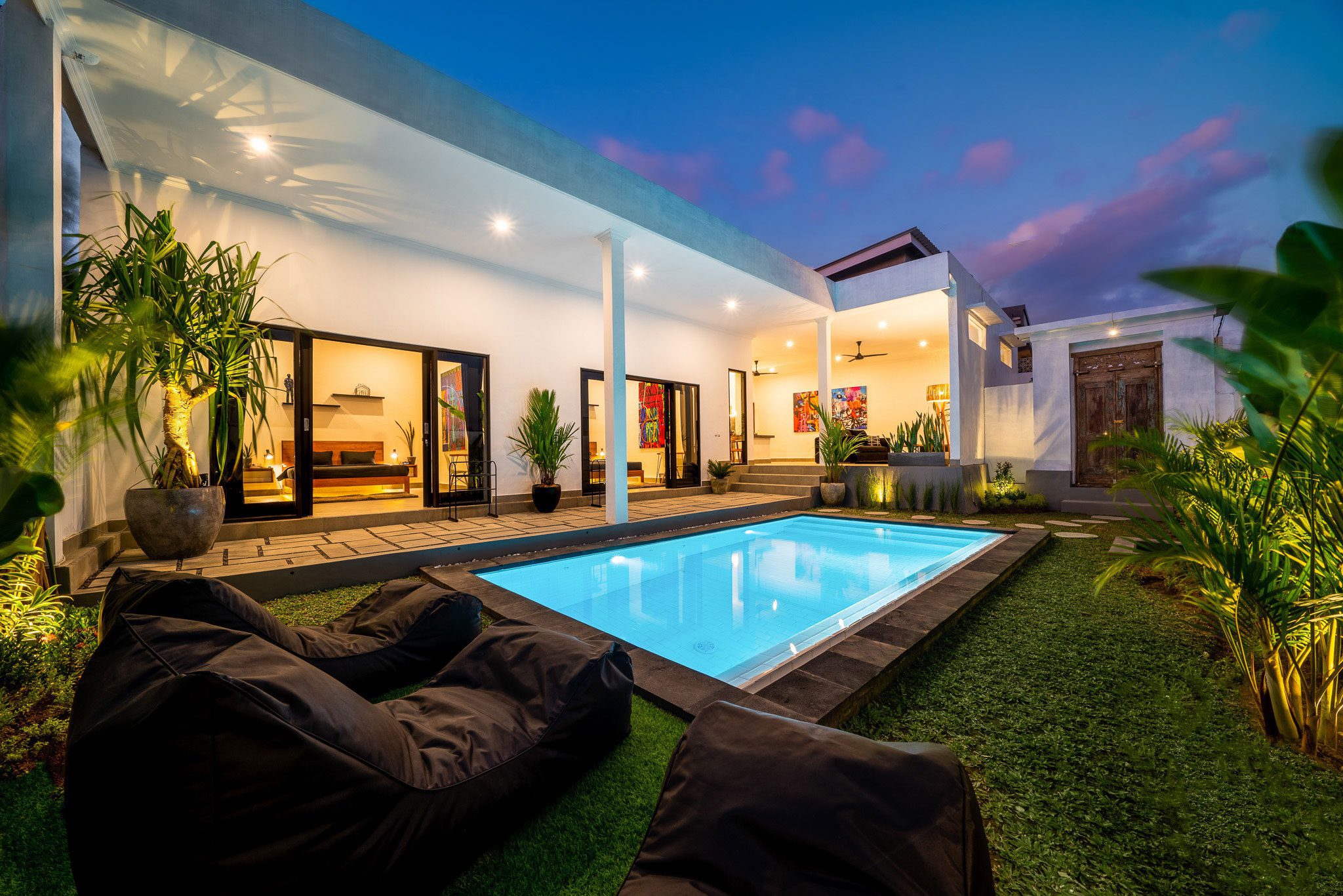 shutterstock_2341050169 Outdoor view of a modern house with a lit rectangular pool, surrounded by greenery and outdoor seating, taken during twilight. Glass doors lead to the interior, and the sky has a mixture of blue and pink hues.